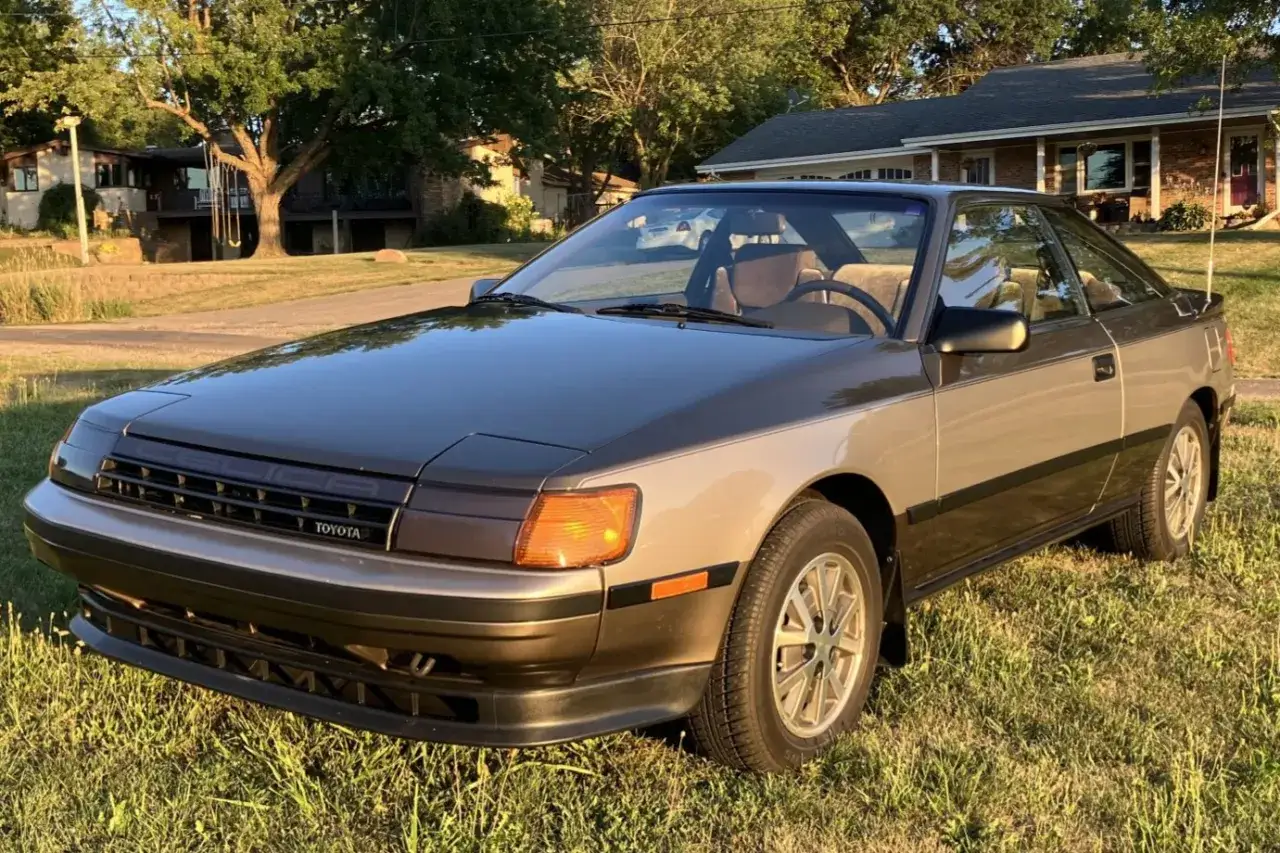 En klassisk Toyota Celica fr&aring;n 1986 st&aring;r parkerad p&aring; en gr&auml;smatta. Bilen har en tv&aring;f&auml;rgad lackering i brunt och gr&aring;tt.