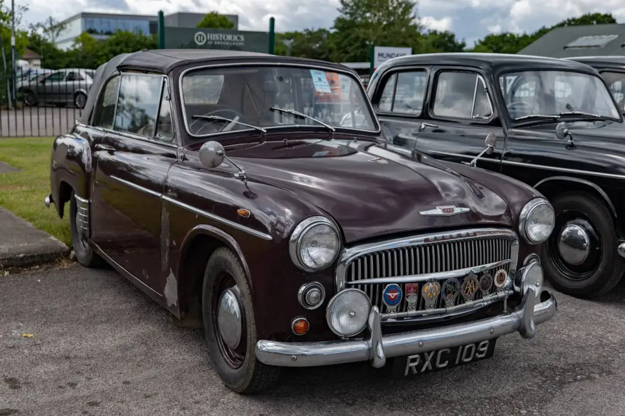 En m&ouml;rklila Hillman-bil med cabriolettak st&aring;r parkerad bredvid en svart bil. Bilen har en kromad grill med flera m&auml;rken.