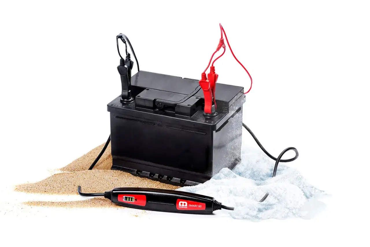 Bilbatteri kopplat till laddare, redo att ladda. Sand och snö runt batteriet symboliserar olika miljöer.