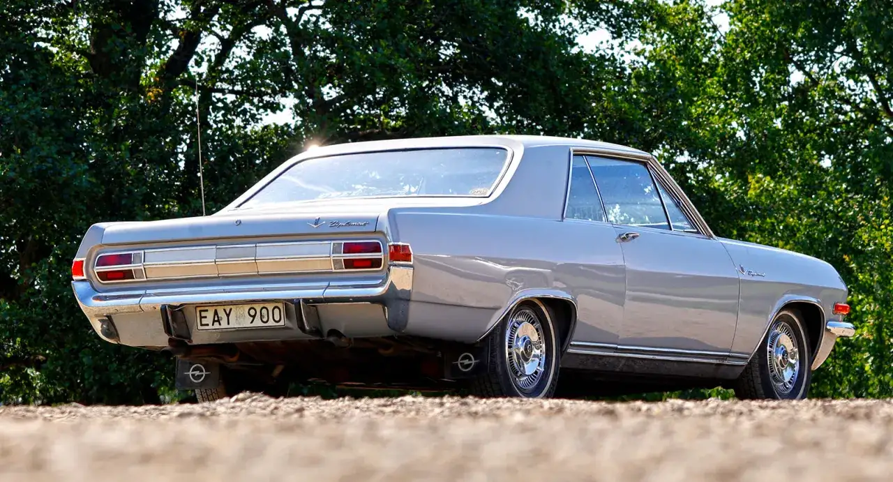 En gr&aring; Opel Diplomat coup&eacute; st&aring;r parkerad utomhus, med en gr&ouml;nskande bakgrund av tr&auml;d.