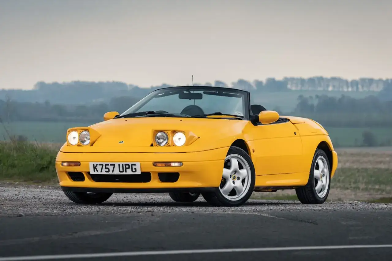 Gul Lotus Elan M100 cabriolet parkerad på grusväg med fält och träd i bakgrunden.
