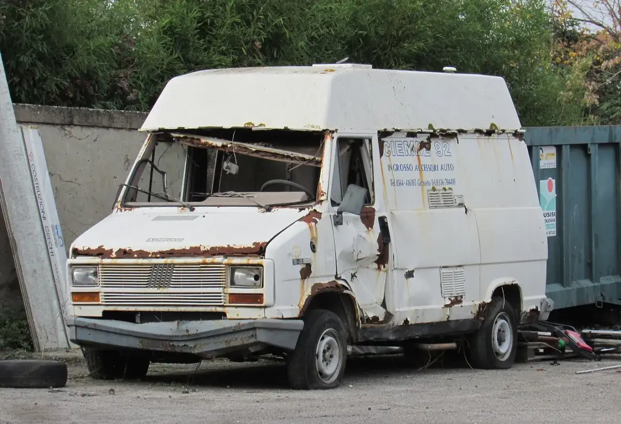 A heavily rusted, white Fiat Ducato van with a broken windshield and dented bodywork.