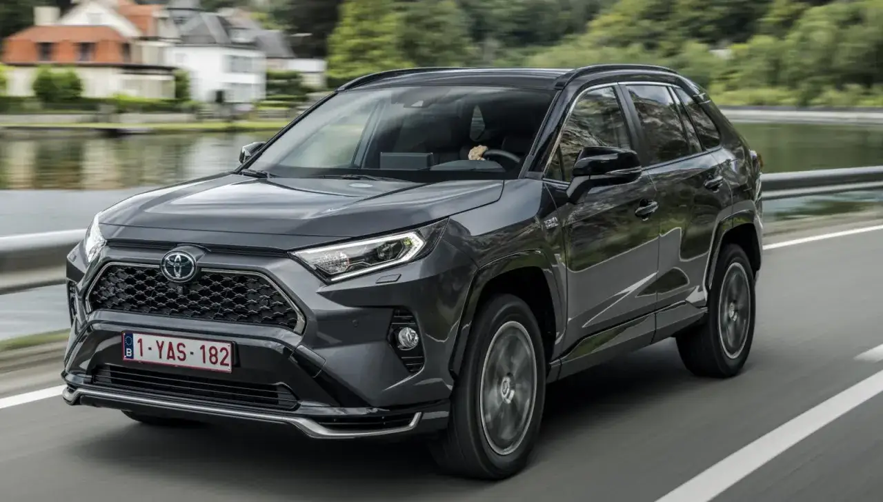 Mörkgrå Toyota RAV4 körs på en väg med en sjö och hus i bakgrunden. En del av en toyota rav4 test.