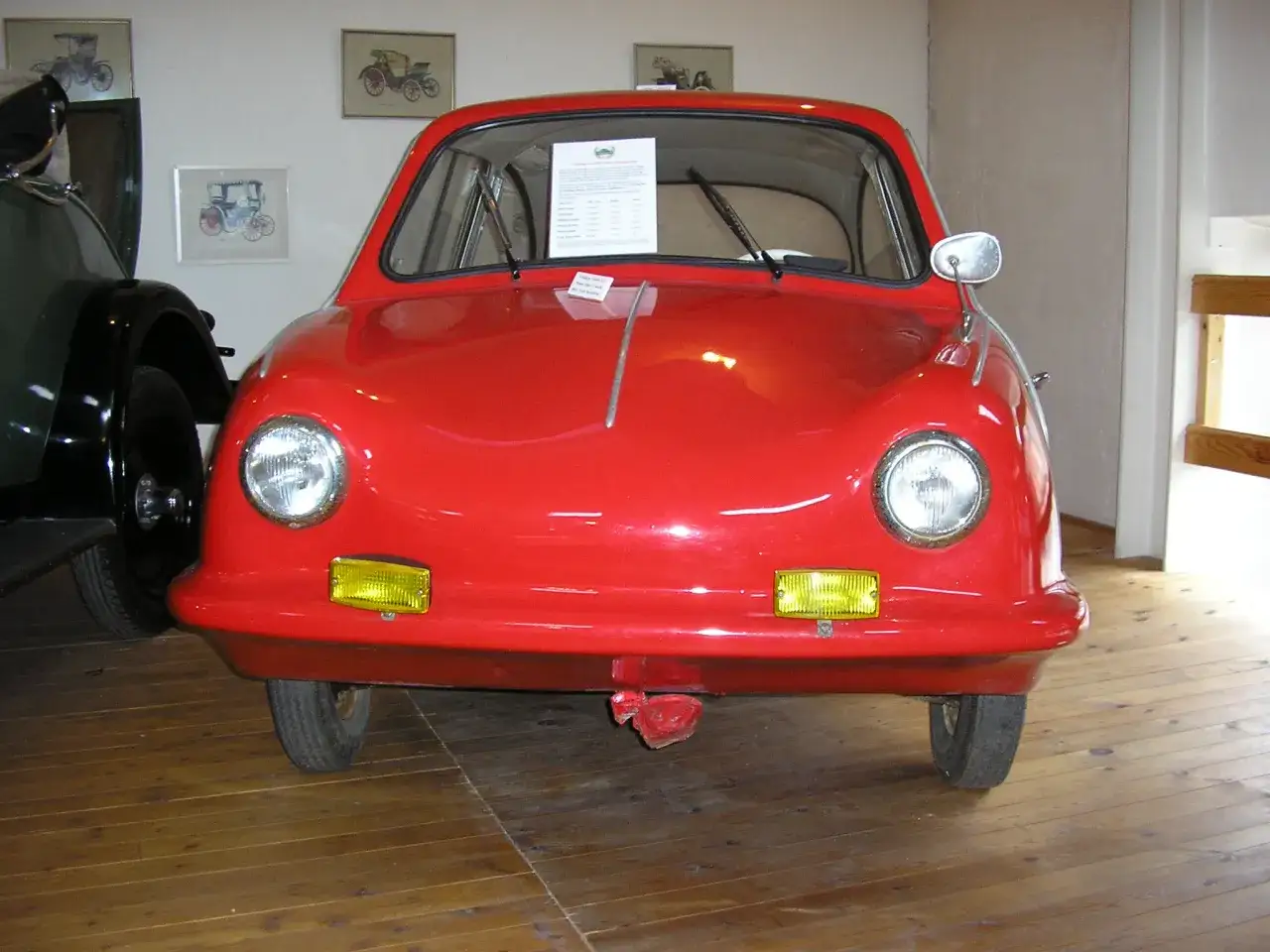 En röd, liten bil, en Messerschmitt KR200, står på ett trägolv. Fram king fulda.