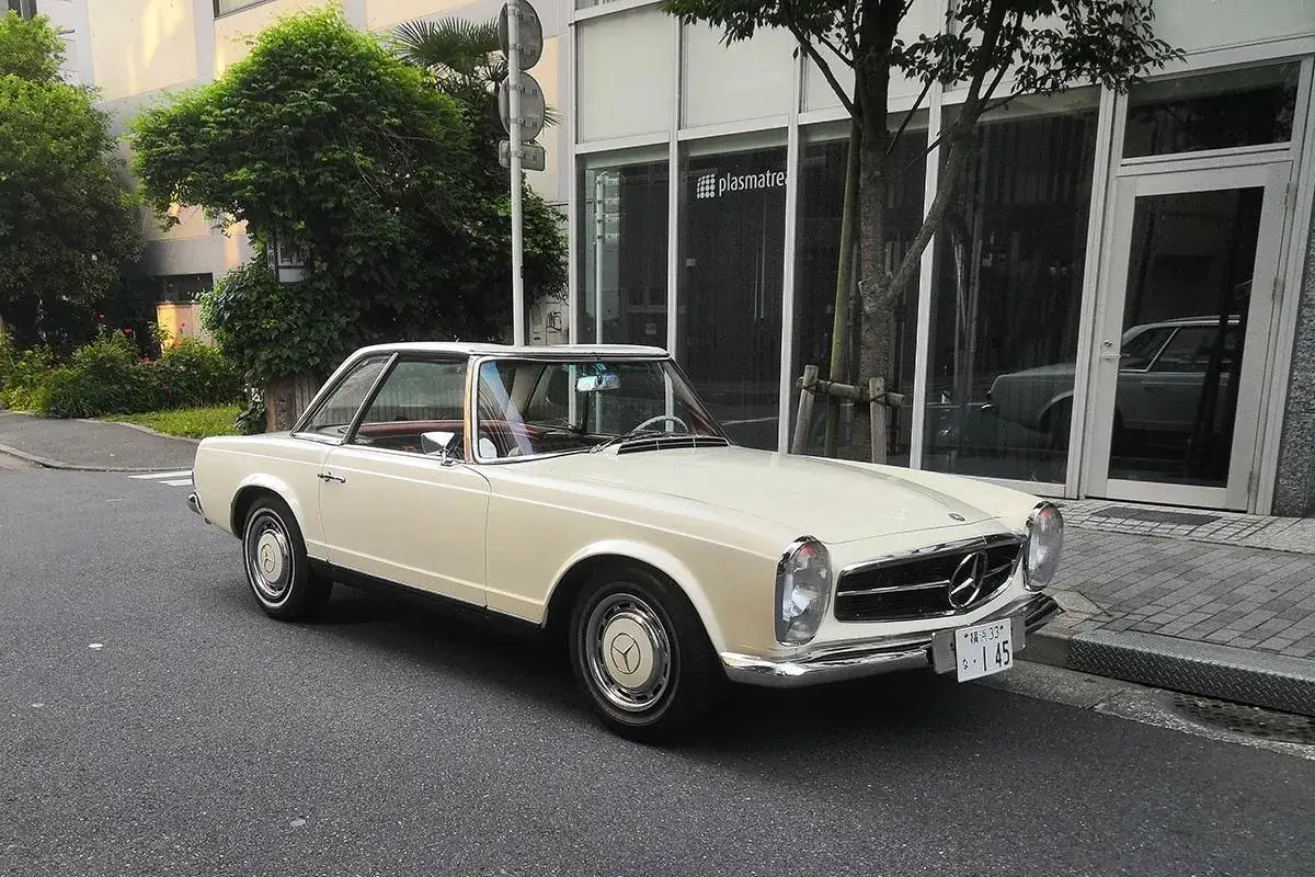 En krämvit Mercedes 230 SL står parkerad på en gata. Bilen har en klassisk design med kromdetaljer.