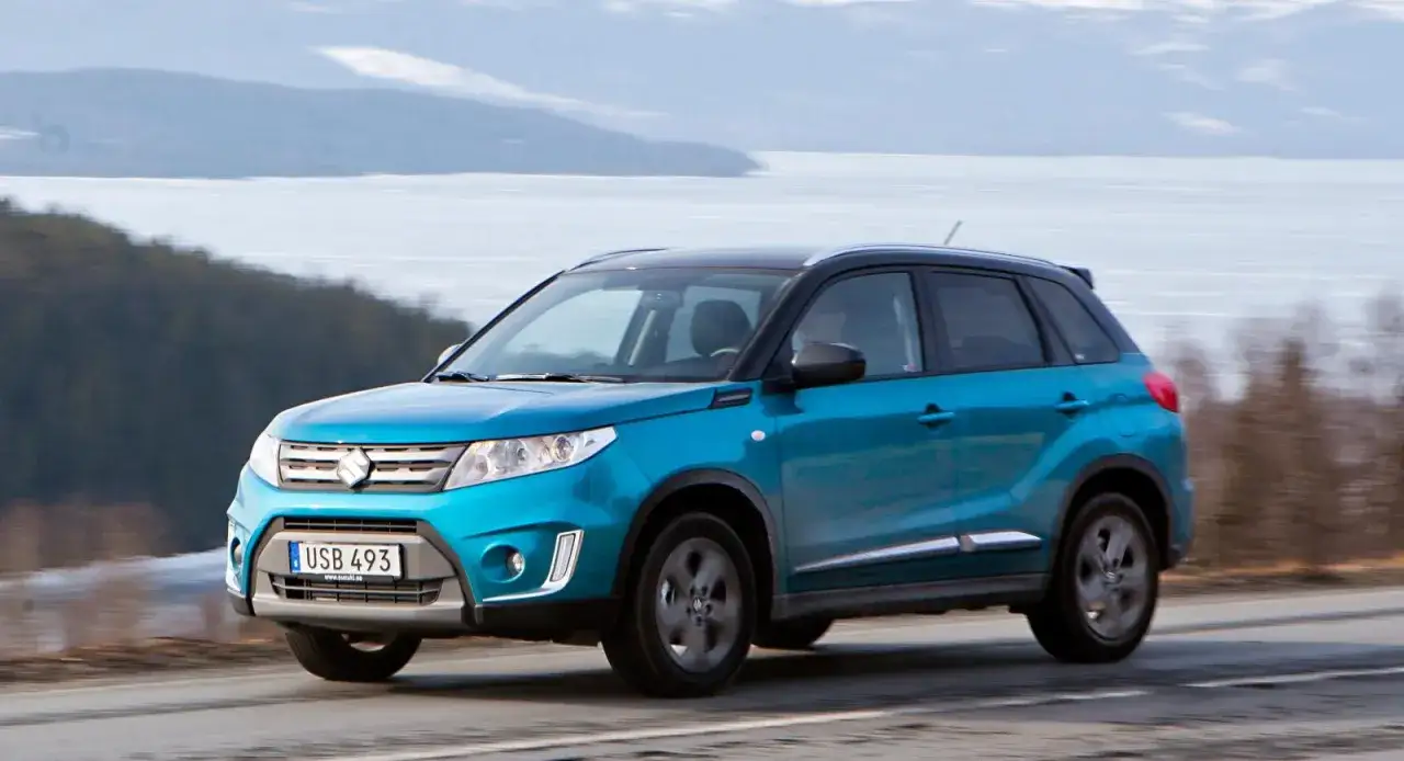Bl&aring; Suzuki Vitara p&aring; v&auml;g l&auml;ngs en v&auml;g med isig sj&ouml; i bakgrunden. En perfekt bil f&ouml;r ett suzuki vitara test i vinterlandskap.