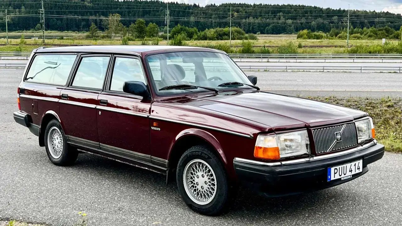 En vinröd Volvo 245 kombi står parkerad vid en väg med skog i bakgrunden.