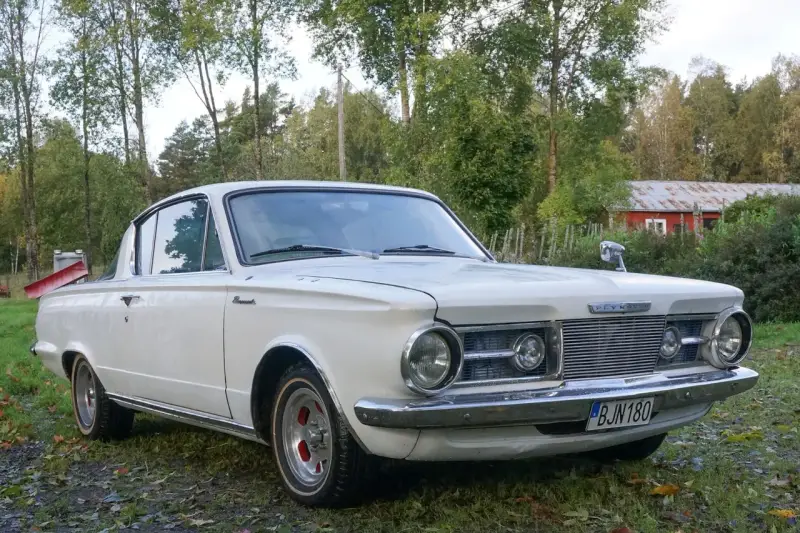 Vit Plymouth Barracuda från 1960-talet parkerad på en gräsmatta med träd och en röd byggnad i bakgrunden.
