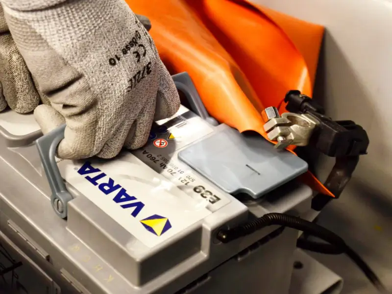 Hand med skyddshandske kopplar ihop kablar för att ladda bilbatteri. Orange skyddsdyna syns bakom.